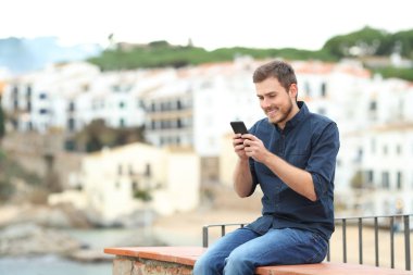 Sahilde tatilde bir çıkıntıda oturmuş akıllı telefondan mesaj atan mutlu bir adam.