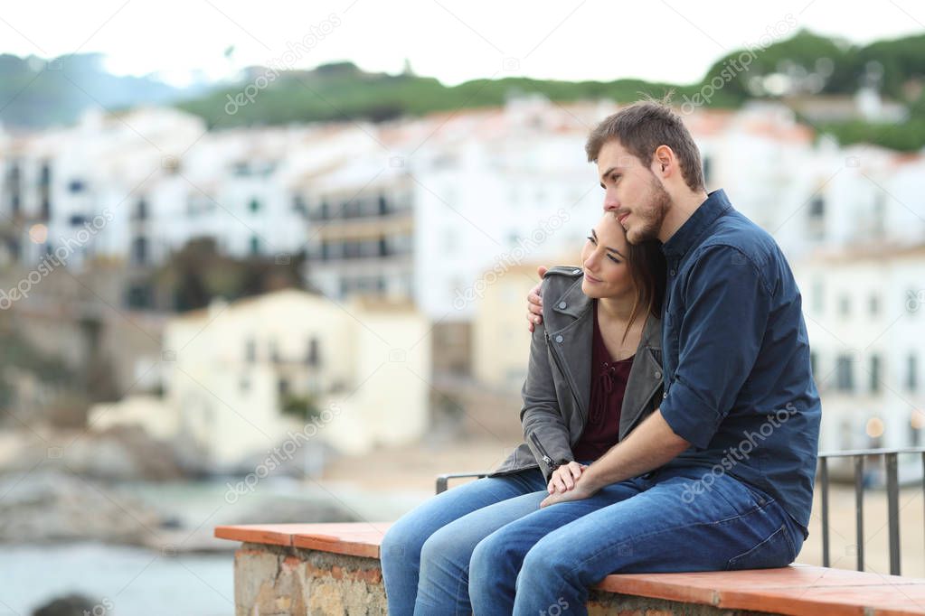 Happy couple in love looking away sitting on a ledge on vacation in a coast town