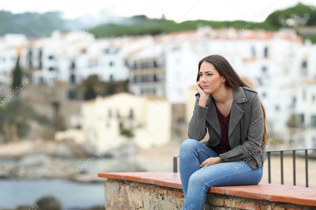 Sad pensive woman looking away sitting on a ledge in a coast town