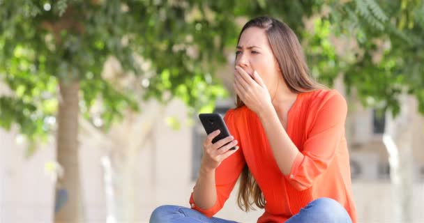 女性あくびを公園のベンチに座っているスマート フォンを使用して — ストック動画