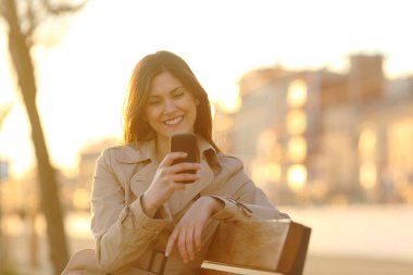 Mutlu kız günbatımı bir parkta bir bankta günbatımında oturmuş., akıllı telefon kontrol