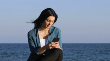 Sahilde bir bankta oturmuş akıllı telefon üzerinde ciddi kadın manifatura