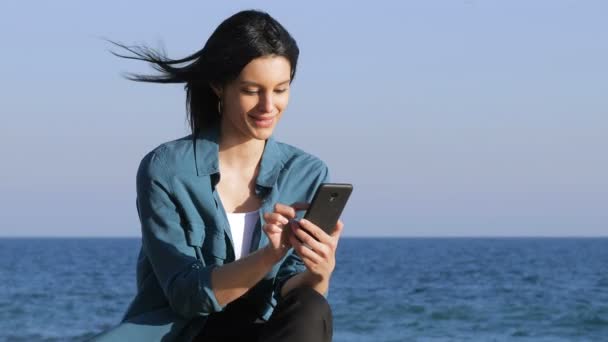 严肃的妇女坐在海滩上的长凳上浏览智能手机内容 — 图库视频影像