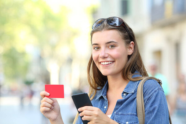 Happy teen holding blank credit or gift card and smart phone looking at camera in the street