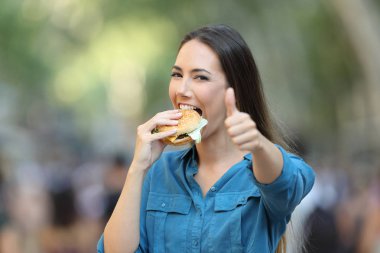 Sokakta hamburger yiyen mutlu bir kadın.