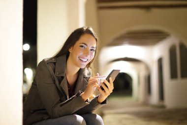 Geceleri akıllı telefon kullanan mutlu kadın sokaktaki kameraya bakıyor.