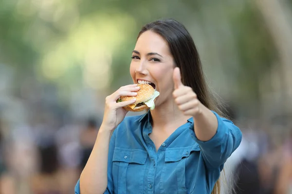 快乐的女人在街上吃汉堡包 摆弄着大拇指 — 图库照片