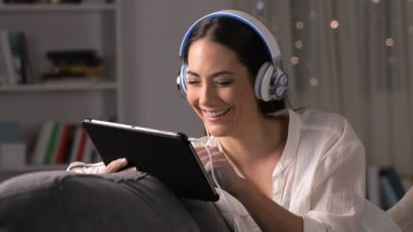 Gece evde bir kanepede oturan tablet medyada izlerken kulaklıklar giyen mutlu kız