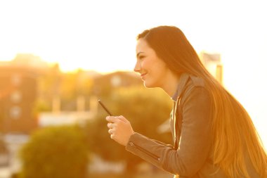 Akıllı telefon uzakta bir balkonda batımını tutan dalgın bir kızın profil