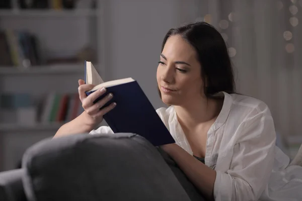 Serious lady reads a book sitting on a couch in the night at home