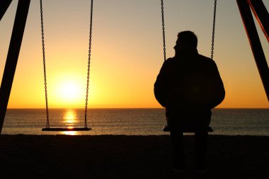 Back view backlighting silhouette of a man alone on a swing looking at empty seat at sunset on the beach in winter clipart