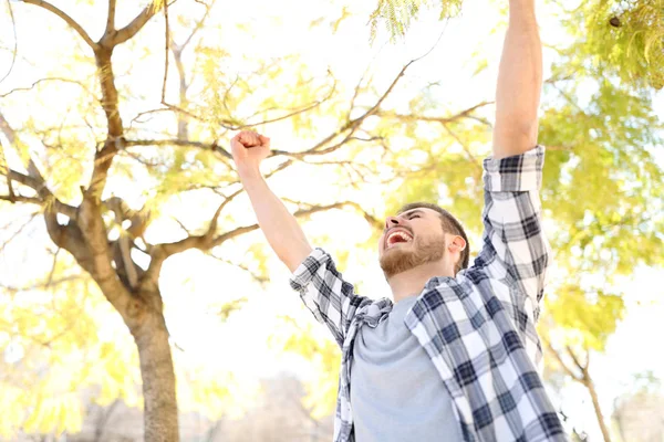 Excited guy celebrating success raising arms in a park with trees in the background