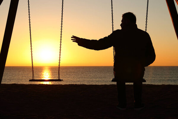 Back view backlighting silhouette of a man sitting on swing alone missing her partner at sunset on the beach in winter