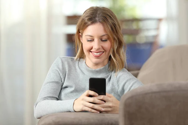 Senhora feliz mensagens de texto no telefone em um sofá em casa — Fotografia de Stock