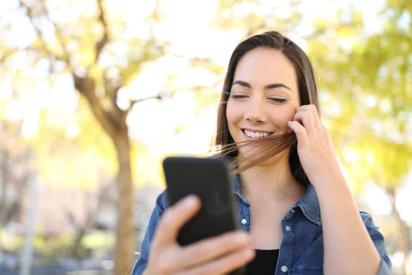 Donna felice utilizzando smart phone toccare i capelli in un parco — Foto Stock