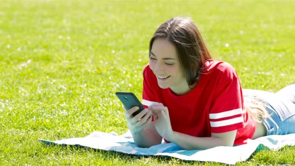 愉快的少女在红色检查电话躺在草地上在公园 — 图库视频影像