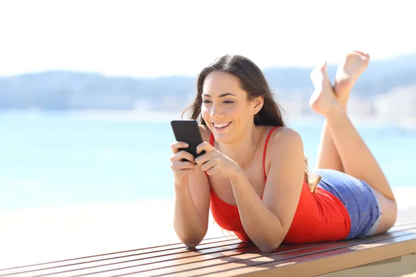 Feliz adolescente usando el teléfono tumbado en un banco en la playa — Foto de Stock