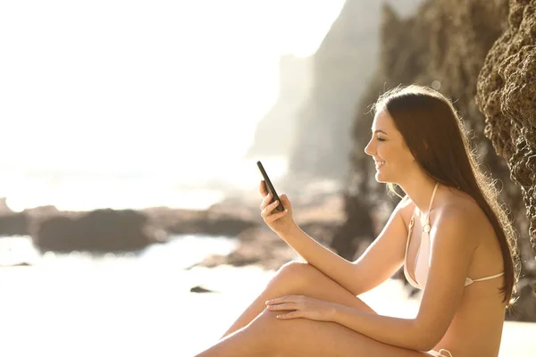 Turista feliz en bikini comprobando teléfono en la playa —  Fotos de Stock