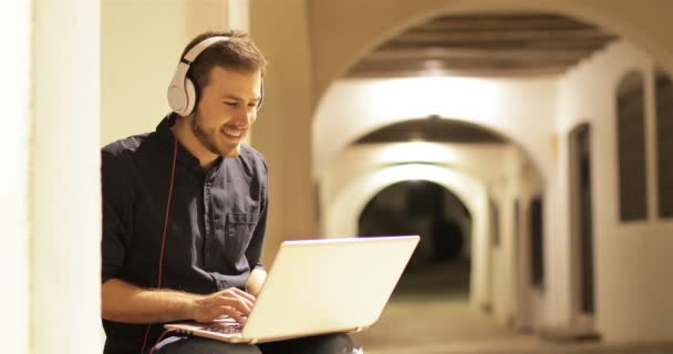 晚上坐在镇上的街道上 快乐的男人使用带耳机的笔记本电脑 — 图库视频影像