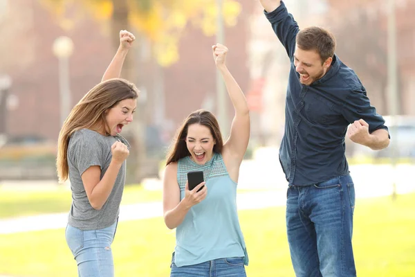 Három izgatott barátok jumping ellenőrzése okos telefon a parkban — Stock Fotó