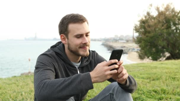 海岸の都市の郊外の草の上に座ってスマートフォン上の幸せな十代のテキストメッセージ — ストック動画