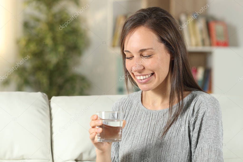 Happy woman holding a glass of water on a couch at home