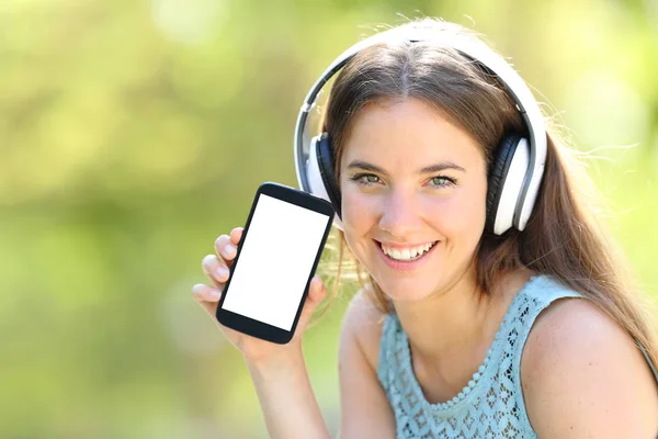 Menina com fones de ouvido mostrando tela de telefone inteligente em branco no verde — Fotografia de Stock