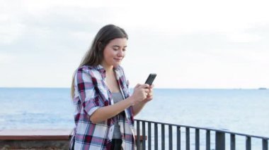 Mutlu genç bir sahil kasabası sokakta kameraya doğru yürüyen bir akıllı telefon kullanarak