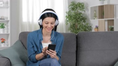 Mutlu kadın indirme ve evde bir kanepede oturan akıllı telefondan müzik dinleme