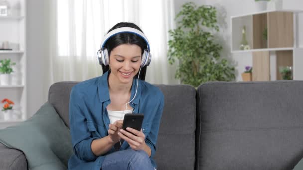 自宅のソファに座ってスマートフォンから音楽をダウンロードして聴く幸せな女性 — ストック動画
