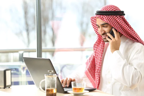 Gelukkige Arabische man met behulp van een laptop en praten op telefoon in een bar — Stockfoto