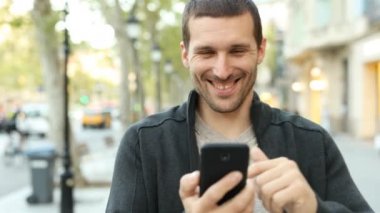Caddede kameraya doğru yürüyen akıllı telefonu kullanan mutlu bir adamın ön görüntüsü.