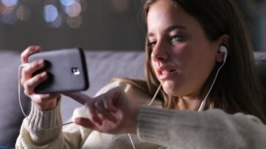 Kulaklıklı bir kadın cep telefonuyla medyayı izliyor. Geceleri koltukta oturuyor. 