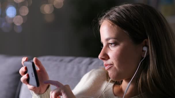 夜にソファに座っている携帯電話でメディアを見てイヤホンを持つ女性 — ストック動画