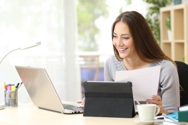 Dizüstü bilgisayarı ve tableti olan mutlu kadın, bir masaevinde oturan belgeyi karşılaştırıyor