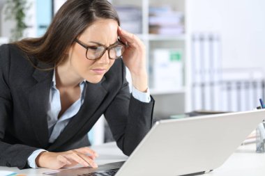 Gözlüklü, gözlüklü bir kadın ofisteki masasında oturmuş laptopta okumaya çalışıyor.