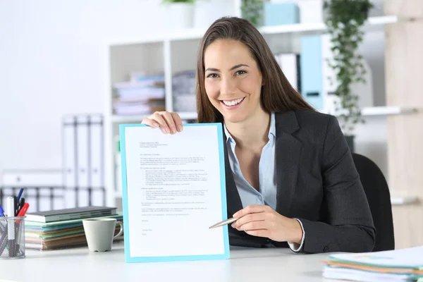 Front view of a fappy executive woman points contract signature space with pen looking at camera sitting on a desk at office