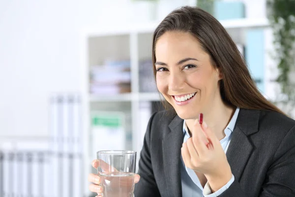 Happy executive woman showing pill with water glass looking at camera sitting on a desk at office