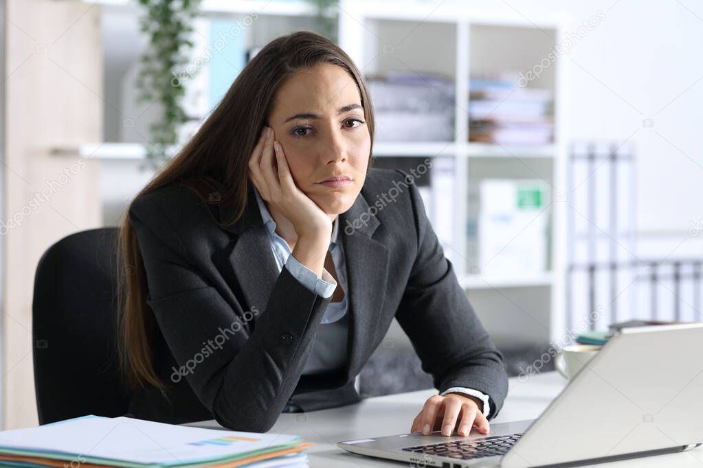 Bored executive woman with laptop looking at camera sitting on her desk at office