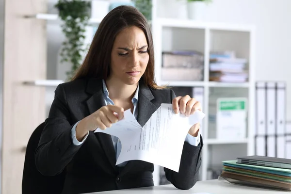 Angry executive woman breaking contract document sitting on a desk in the office at night