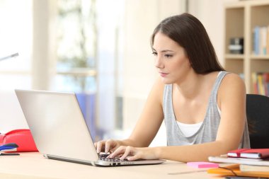 Evdeki bir masada oturup dizüstü bilgisayarda yazan ve okuyan ciddi bir öğrenci kadın.