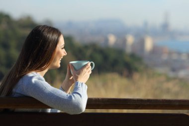 Kenar mahallede bir bankta elinde kahve fincanı tutan mutlu bir kadının yan görüntüsü.