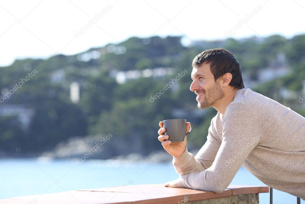 Side view of a happy man contemplating views with coffee cup on a balcony on the beach