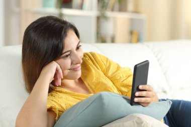 Akıllı telefondan medyayı izleyen bir kadın evdeki oturma odasındaki kanepede uzanıyor.