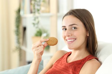 Mısır gevreği kurabiyesi tutan mutlu kadın evdeki oturma odasında bir kanepede otururken sana bakıyor.