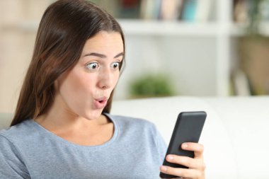 Şaşırmış kız, evdeki oturma odasındaki koltukta oturan akıllı telefonu kontrol ediyor.