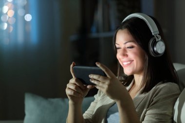Kulaklık takan mutlu bir kadın gecenin bir yarısı akıllı telefondan video izleyip evdeki oturma odasında bir kanepede oturuyor.