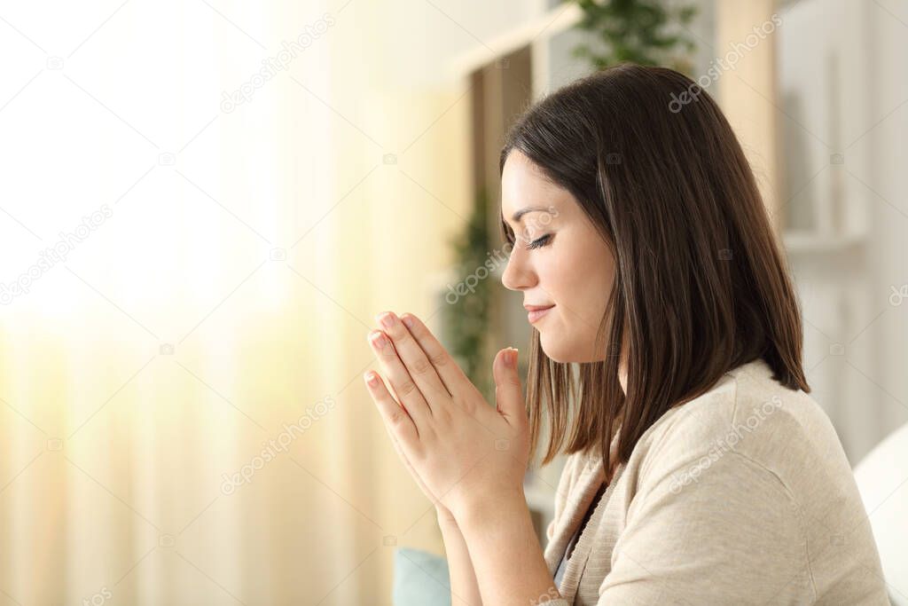 Side view of a christian religious woman praying sitting on a couch in the living room at home with window light in the background