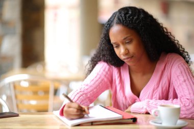 Kafe terasında notlar alan konsantre siyah öğrenci.