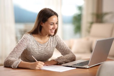 Evdeki ahşap masada dizüstü bilgisayarı kontrol eden mutlu bir kadın.